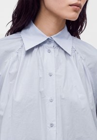 Femme portant une chemise ample bleu clair à boutons avec un large col, montrée de la poitrine au menton sur un fond blanc.