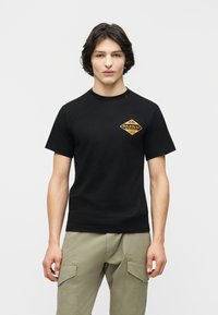 Νεαρός άνδρας με μεσαίου μήκους σκούρα μαλλιά, που φοράει μαύρο μπλουζάκι με κίτρινο λογότυπο Miami Replay Palace Florida και μπεζ παντελόνι καμπαρντίνα.