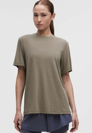 Vrouw met bruin haar naar achteren gebonden, die een olijfgroen T-shirt met korte mouwen en donkerblauwe korte broek draagt tegen een effen achtergrond.