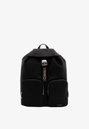 Sac à dos en cuir noir Calvin Klein avec fermeture à rabat, sangle avant avec boucle et deux poches avant zippées sur fond blanc.