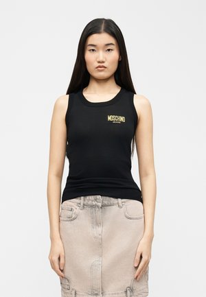 Νεαρή γυναίκα με μακριά σκούρα μαλλιά φοράει μαύρη αμάνικη μπλούζα με κίτρινο λογότυπο «Moschino Jeans» και φούστα τζιν ανοιχτού μπεζ χρώματος.