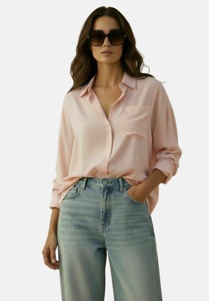 Vrouw met lang bruin haar die oversized zwarte zonnebrillen draagt, een lichtroze blouse met knopen en lichtblauwe jeans met hoge taille.