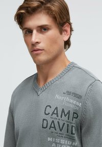 Grauer V-Ausschnitt-Pullover mit geripptem Kragen, strukturierter Stoff und bedruckte Grafiken mit "CAMP DAVID" in fetten schwarzen Buchstaben.