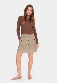 Femme portant un haut marron à manches longues, une jupe beige à boutons devant avec poches, et des ballerines beige à clous, se tenant les mains dans les poches sur fond blanc.