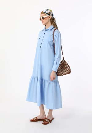 Light blue shirt dress with long sleeves, two chest pockets, and a tiered hem. Paired with brown sandals and a woven brown handbag.