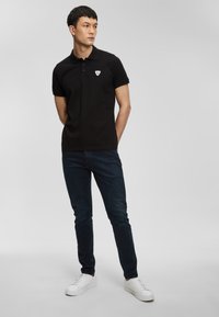 Černé polo tričko s límečkem, třemi knoflíky a malým trojúhelníkovým logem. Nosené s tmavými džínami a bílými teniskami.