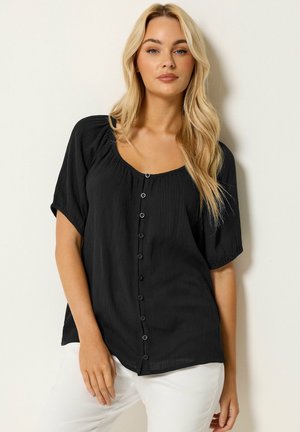 Blonde Frau trägt eine schwarze kurzärmelige, geknöpfte Bluse und weiße Hose, steht vor einem einfachen hellen Hintergrund.