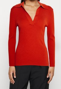 Czerwony prążkowany sweter dziany z dekoltem w kształcie litery V i kołnierzykiem, z długimi rękawami i dopasowanym krojem. Teksturowana tkanina tworzy gładkie wykończenie.