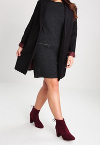 Manteau noir avec poignets bordeaux, robe en maille grise avec détail zippé, et bottines en daim bordeaux avec un accent noué.