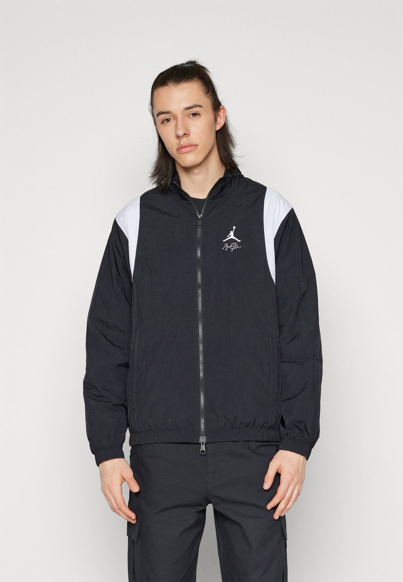 Veste coupe-vent noire avec accents blancs sur les épaules, fermeture éclair à l'avant et un petit logo. Comprend des poignets et un ourlet élastiques. Tissu léger.