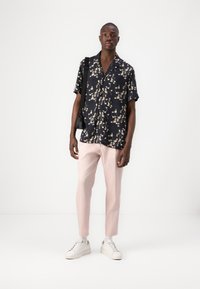 Kortärmad svart skjorta med blommönster, rosa avsmalnande byxor och vita sneakers. Modellen håller en liten svart väska och står framför en vit bakgrund.