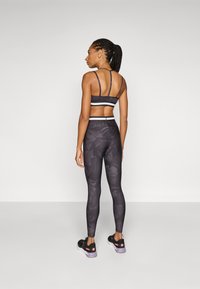 Fekete sportmelltartó vékony pántokkal, fehér díszítéssel, és passzoló fekete kamuflázs leggings. Fekete sportcipővel viselve, amelyen rózsaszín részletek találhatók.