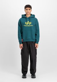 Sudadera verde azulado con un logo y texto amarillos, bolsillo estilo canguro, capucha con cordón, combinada con pantalones cargo negros y zapatillas negras.