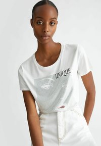 T-shirt bianco in cotone con collo a giro, caratterizzato da una stampa grafica in rete con il testo "UNIQUE" e dettagli rossi. Abbinato a una gonna bianca.