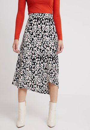 Femme portant une jupe midi à motif noir et blanc, un haut à manches longues orange et des bottines blanches, sur un fond clair uni.