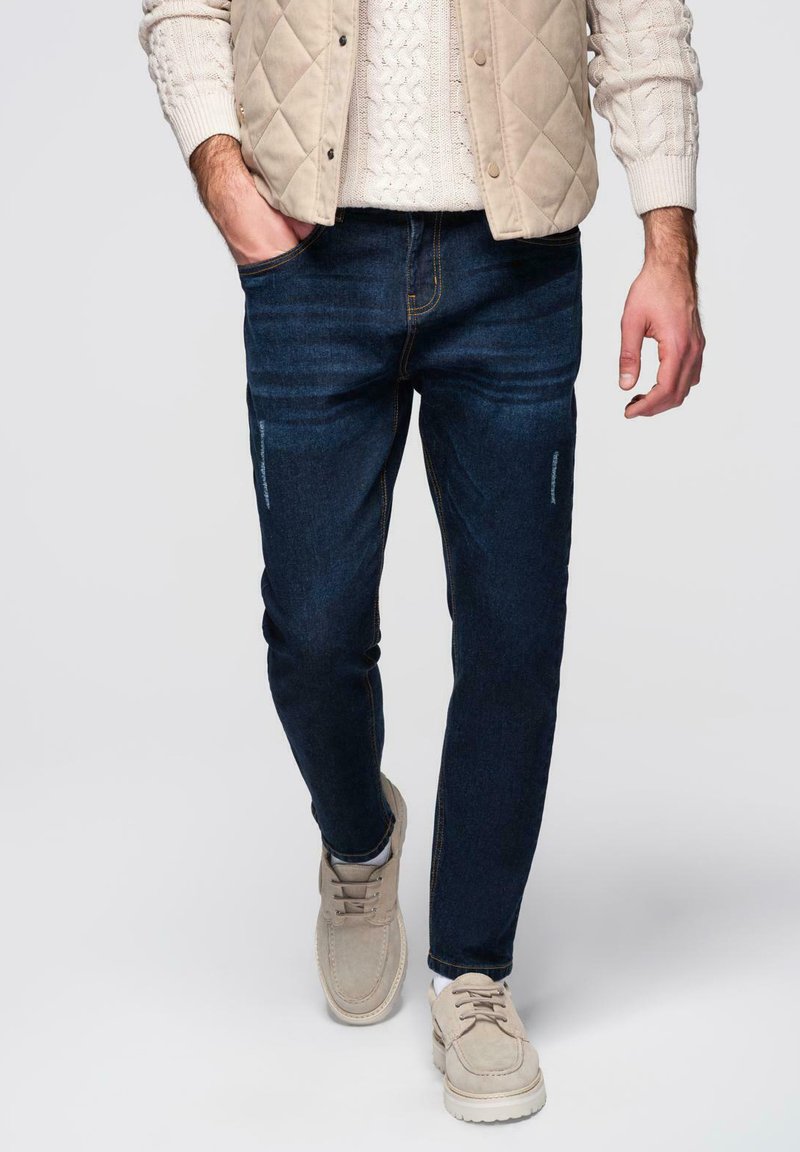 Jeans bleu foncé à coupe slim, design à cinq poches avec un léger effet délavé. Associés à un pull crème en maille torsadée et des chaussures beiges.