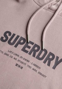 Lichtroze hoodie van zachte stof met een groot zwart "SUPERDRY" logo en kleine gedrukte details nabij de zoom en de trekkoorden.