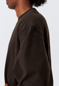 Brauner Baumwollpullover mit Rundhalsausschnitt, überschnittenen Schulternähten und einer weichen Textur, von der Seite gezeigt.