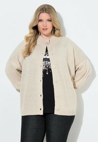 Cardigan oversize beige chiaro con scollo rotondo, chiusura a bottoni e orlo a coste. Indossato sopra una camicia nera con stampa animalier. Tessuto morbido.