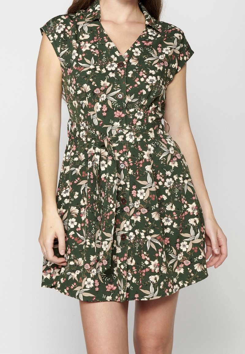 Femme portant une robe courte fleurie vert foncé avec des boutons sur le devant et une ceinture en tissu assortie nouée à la taille.