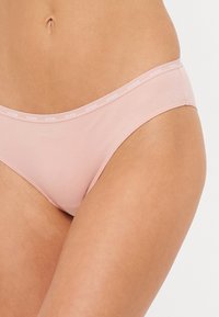Light pink cotton bikini underwear with elastic waistband featuring repeating "GUESS" logo. Smooth texture, minimal coverage design.