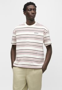 Άνδρας που φοράει λευκό T-shirt με λεπτές οριζόντιες ροζ και καφέ ρίγες και μπεζ παντελόνι, στέκεται με τα χέρια στις τσέπες.