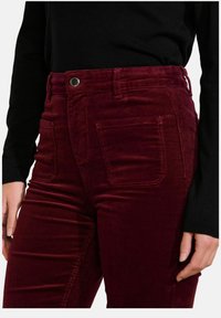 Pantalones de pana burdeos de talle alto con bolsillos frontales visibles y costuras de contraste; combinados con una blusa negra de manga larga.