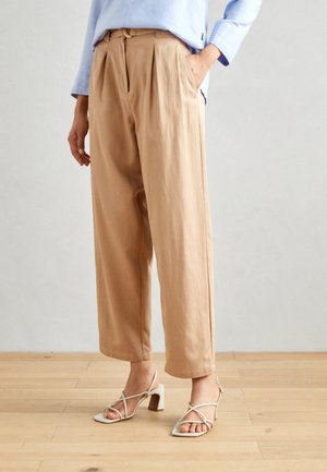 Pantalons plissés beiges avec poches latérales, taille haute et coupe décontractée. Associés à des sandales à talons blancs à lanières.