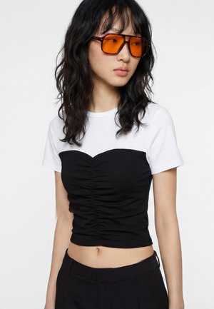 Giovane donna con capelli neri ondulati che indossa occhiali da sole con lenti arancioni, una camicia bianca sovrapposta a un top nero arricciato e pantaloni neri.