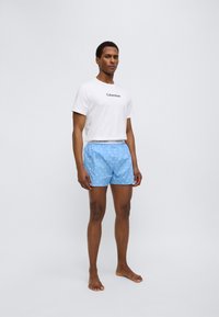 Ανδρικό ντύσιμο με λευκό βαμβακερό t-shirt με μαύρο λογότυπο και ανοιχτό μπλε σορτς με λευκό σχέδιο, σχεδιασμένο για άνεση και casual εμφάνιση.