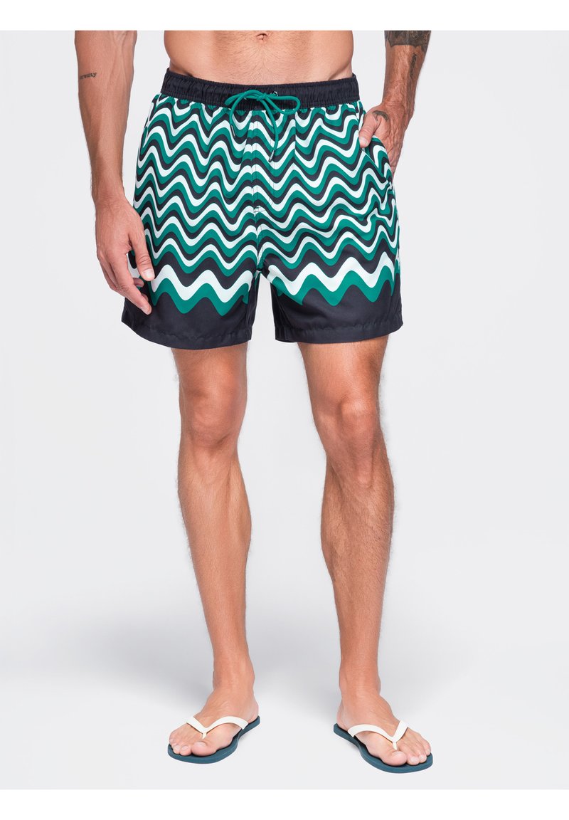 Homme portant un short de bain vert et noir à motif ondulé et des tongs blanches, debout avec une main dans la poche.