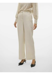 Pantalons beige à jambe large avec une texture lisse, dotés d'une taille haute et de poches avant, associés à des escarpins noirs à bout pointu.