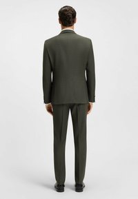 Dunkelgrüner Anzug mit strukturiertem Design, aus strukturiertem Stoff gefertigt. Verfügt über einen Schlitz auf der Rückseite, passende Hose und geschlossene schwarze Schuhe.