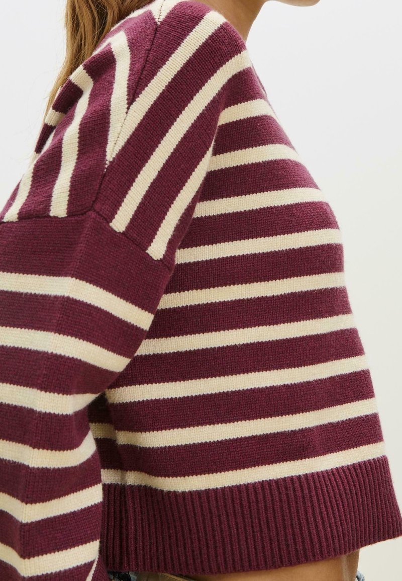 Femme portant un pull tricoté court à rayures bordeaux et crème avec des manches longues, sur un fond uni.