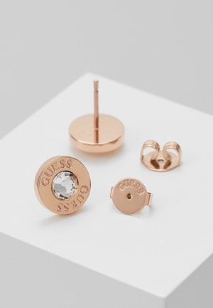 Boucles d’oreilles clous en or rose avec pierres précieuses transparentes, gravées "GUESS", présentées avec attaches assorties sur une surface blanche.