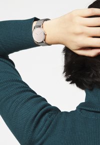 Silberne Armbanduhr mit rundem Zifferblatt, Netzarmband und minimalistischem Design. Die Hand befindet sich in der Nähe eines dunkelblauen, gerippten Pullovers.