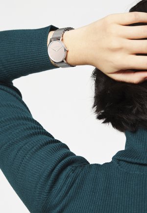 Montre en argent avec un cadran rond, un bracelet en maille et un design minimaliste. Main positionnée près d'un pull côtelé bleu canard sombre.