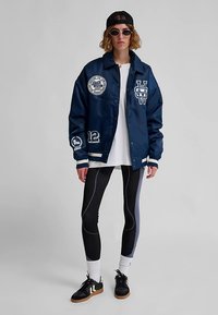 Navy varsity jacket with embroidered patches, white stripes, paired with black and gray leggings, white socks, and black sneakers.