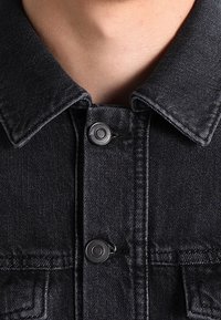 Svart denimjacka med en klassisk krage, framknappar med tryckknappar och bröstfickor med synlig sömdetalj. Texturerat tyg.