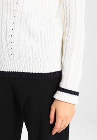 Biały sweter w sploty z dzianiny, z prążkowanym granatowym wykończeniem na dole i na rękawach, o teksturowanym wzorze i dopasowanym kroju.