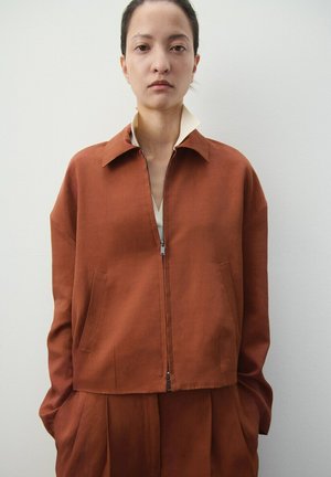 Femme debout contre un mur uni portant une veste zippée couleur rouille sur un pantalon plissé assorti, les mains dans les poches.