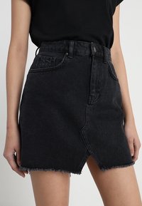 Gonna di denim nera con orlo sfilacciato, design a vita alta e spacco frontale. Presenta una tasca singola e dettagli cuciti.