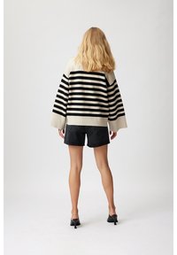 Mujer con cabello rubio que lleva un suéter a rayas blanco y negro, pantalones cortos de mezclilla negros y mule de tacón negro, de pie contra un fondo liso.
