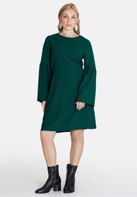 Rochie verde cu maneci lungi, decolteu rotund, maneci evazate și talie ajustată. Purtați cu bocanci negri până la gleznă. Textură de țesătură netedă.