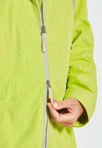 Helle grüne Jacke mit einem Reißverschluss über die gesamte Länge, aus strukturiertem Stoff und einer Hand, die den Reißverschlussgriff hält. Einfacher Tascheneingriff auf der Vorderseite.