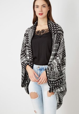 Joven mujer con una blusa negra con encaje, jeans claros rasgados y un chal con estampado blanco y negro sobre un fondo liso.