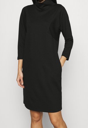 Femme portant une robe noire mi-longue à manches longues avec un col roulé ample, debout avec une main dans la poche.