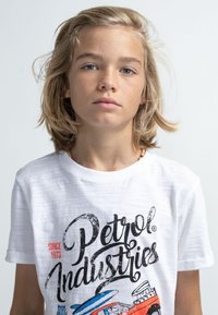Bílý tričko s krátkým rukávem s grafickým potiskem barevného auta a stylizovaným textem "Petrol Industries" s červenými akcenty.