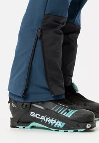 Zwarte en blauwe skibroek met ritssluiting aan de zijkant en elastische manchetten, gecombineerd met een zwarte skiboot met turquoise accenten en merkdetails.