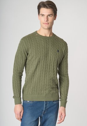 Giovane uomo con maglione verde a trecce e jeans blu, in piedi contro uno sfondo chiaro uniforme, leggermente rivolto verso destra.